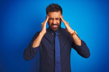 Zarif gömlek ve kravat giyen genç Hintli işadamı, eliyle baş ağrısı nedeniyle stres ekibe sahip izole mavi arka plan üzerinde duruyor. Acı çeken migren.