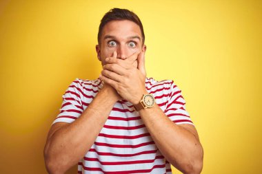 Genç yakışıklı adam sarı izole arka plan üzerinde rahat kırmızı çizgili t-shirt giyen hata için elleri ile ağız kapsayan şok. Gizli kavram.