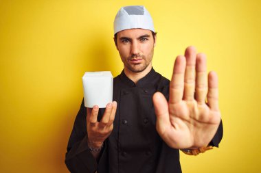 Üniforma ve şapka tutan teslimat kutusu giyen genç şef, izole sarı arka plan üzerinde açık el ile ciddi ve kendinden emin ifade, savunma hareketi ile dur işareti yapıyor