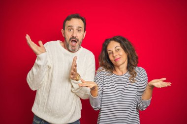 Beautiful middle age couple wearing winter sweater over isolated red background clueless and confused expression with arms and hands raised. Doubt concept.