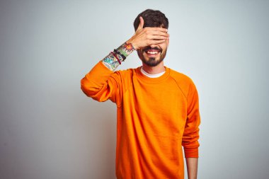 Dövmegiyen turuncu kazak giyen genç adam izole beyaz arka plan üzerinde duran gülümseyerek ve sürpriz için gözleri kaplayan yüzünde el ile gülüyor. Kör kavramı.