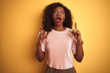 Pembe t-shirt giyen Afro-Amerikalı bir kadın izole edilmiş sarı arka planda durup şaşkın şaşkın yukarı bakıyor ve parmaklarıyla işaret ediyor ve kollarını kaldırıyordu..