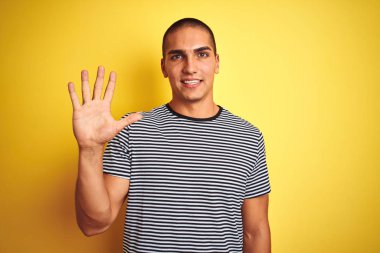 Sarı arka plan üzerine çizgili tişört giyen genç yakışıklı adam beş numaralı parmaklarını gösterip gülümserken kendinden emin ve mutlu görünüyor..