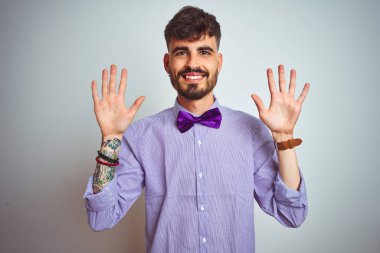 Dövme mor gömlek ve izole beyaz arka plan üzerinde papyon giyen genç adam gösteren ve emin ve mutlu gülümserken parmakları on numara ile işaret.