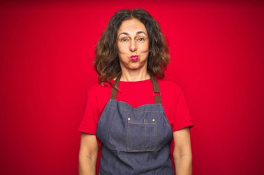 Orta yaş yaşlı kadın komik yüzü ile kırmızı izole arka plan şişme yanakları üzerinde önlük üniforma giyiyor. Ağız hava ile şişirilmiş, deli ifade.