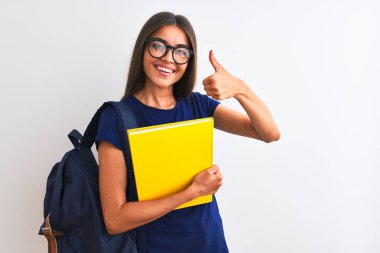 Sırt çantası gözlüğü takan genç bir bayan izole edilmiş beyaz arka plan üzerine kitap tutuyor. Kocaman gülümsüyor. Tamam işareti yapıyor.