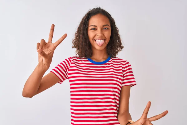 赤のストライプのTシャツを着た若いブラジル人女性が 両手の指が勝利サインをしているのを見て 舌で微笑んだ孤立した白い背景の上に立っています 第二番 — ストック写真