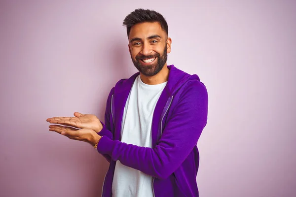 孤立したピンクの背景の上に立つ紫色のスウェットシャツを着た若いインド人男性が手を開けて手のひらを開き 笑顔で幸せで自信を持って広告を提示 — ストック写真