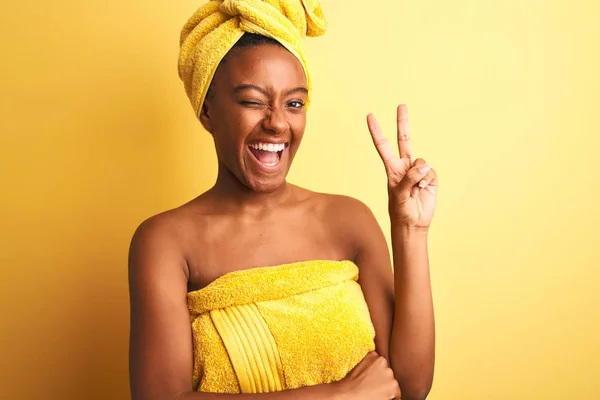 African american woman wearing towel after shower standing over isolated yellow background smiling with happy face winking at the camera doing victory sign. Number two.