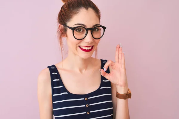 Redhead woman with pigtail wearing striped t-shirt standing over isolated pink background doing ok sign with fingers, excellent symbol