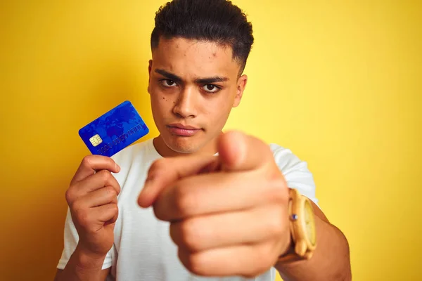 Young brazilian customer man holding credit card standing over isolated yellow background pointing with finger to the camera and to you, hand sign, positive and confident gesture from the front