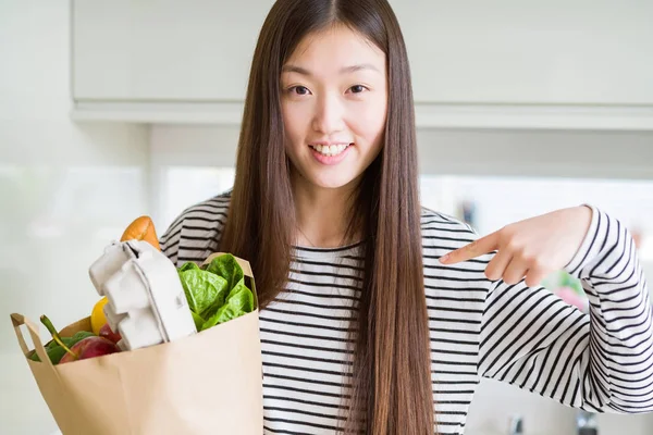 Beautiful Asian woman holding paper bag of fresh groceries with surprise face pointing finger to himself