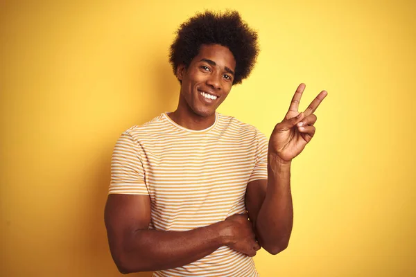 American man with afro hair wearing striped t-shirt standing over isolated yellow background smiling with happy face winking at the camera doing victory sign with fingers. Number two.