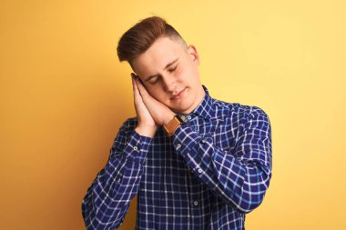 Sarı arka plan üzerinde duran rahat tişört giyen genç yakışıklı adam yorgun uyuyor ve elleri ile birlikte poz verirken kapalı gözlerle gülümsüyor..