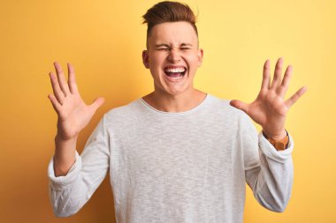 İzole edilmiş sarı arka plan üzerinde duran beyaz tişörtlü genç yakışıklı adam kollarını kaldırmış ve gözleri kapalı heyecanla haykırarak çılgın ve çılgın başarısını kutluyor. Kazanan konsept