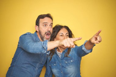 Beautiful middle age couple together standing over isolated yellow background Pointing with finger surprised ahead, open mouth amazed expression, something on the front