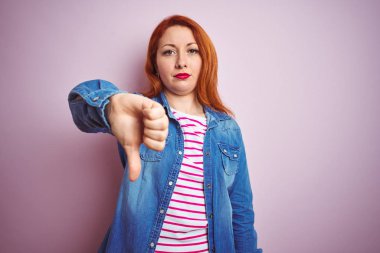 Güzel kızıl saçlı kadın kot gömlek ve pembe arka planda çizgili tişört giyerek mutsuz, kızgın, reddedilmiş ve olumsuz jestler yapıyor. Kötü ifade.
