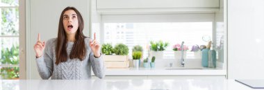 Güzel genç kadın evde beyaz masaya oturan geniş açı resim hayret ve yukarıya bakıp parmakları ile işaret şaşırttı ve silah kaldırdı.