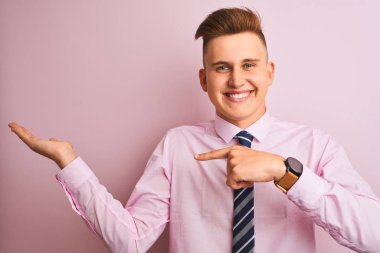 Genç yakışıklı işadamı gömlek ve kravat izole pembe arka plan üzerinde duran giyen hayran ve el ile sunarken kameraya gülümseyerek ve parmak ile işaret.