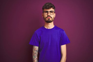 Dövmeli genç adam t-shirt ve gözlük giyen yüzünde ciddi ifade ile izole mor arka plan üzerinde duruyor. Kameraya basit ve doğal bir bakış.