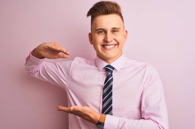 Genç yakışıklı işadamı gömlek ve kravat büyük ve büyük boy işareti gösteren elleri ile izole pembe arka plan gesturing üzerinde duran giyiyor, sembol ölçüsü. Kameraya bakarak gülümsüyor. Ölçme kavramı.