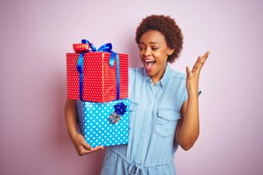 Pembe izole arka plan üzerinde doğum günü hediyeleri tutan Afrikalı Amerikalı kadın çok mutlu ve heyecanlı, kazanan ifade büyük bir gülümseme ile çığlık zafer kutluyor ve ellerini kaldırdı