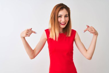 İzole beyaz arka plan üzerinde duran zarif kırmızı elbise giyen redhead iş kadını yüzünde gülümseme ile emin görünümlü, parmakları gururlu ve mutlu kendini işaret.
