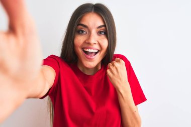 Kırmızı t-shirt giyen güzel bir kadın kamerayla selfie çeker izole edilmiş beyaz arka planda gururla bağırır ve zaferi ve başarıyı kutlar. Çok heyecanlı, coşkulu bir duygu.