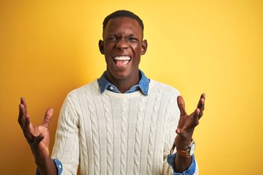 Afro-Amerikan adam denim gömlek ve beyaz kazak giyen izole sarı arka plan üzerinde deli ve deli bağırarak ve agresif ifade ve kollar kaldırdı ile bağırıyor. Hayal kırıklığı kavramı.