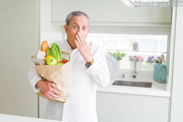 Handsome senior man holding a paper bag of fresh groceries at the kitchen cover mouth with hand shocked with shame for mistake, expression of fear, scared in silence, secret concept