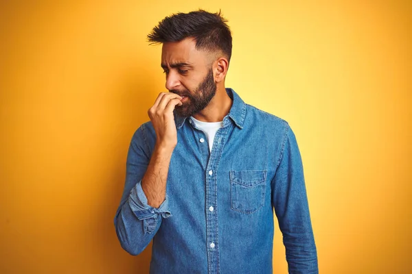 孤立した黄色の背景の上に立つデニムシャツを着た若いインド人男性は 口を噛む爪に手を当て ストレスと緊張しているように見えます 不安の問題 — ストック写真