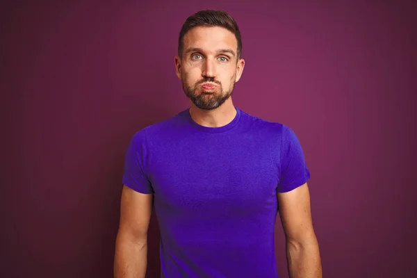 Young man wearing casual purple t-shirt over lilac isolated background puffing cheeks with funny face. Mouth inflated with air, crazy expression.