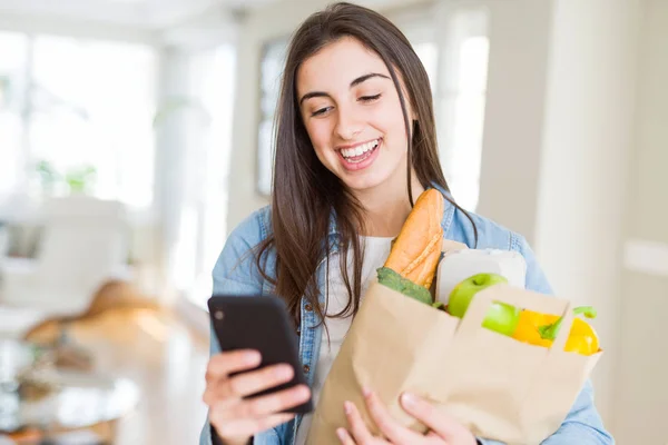 Young woman holding a paper bag full of fresh groceries and usin