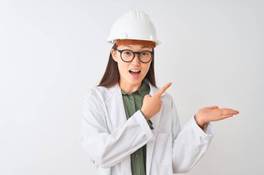 İzole beyaz arka plan üzerinde ceket kask gözlük takan genç Çinli mühendis kadın şaşırttı ve el ile sunarken kameraya gülümseyerek ve parmak ile işaret.