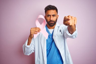 Genç hint doktor adam izole pembe arka plan üzerinde kamera ve size işaret eden üzerinde duran kanser kurdele tutan, el işareti, önden olumlu ve kendinden emin jest