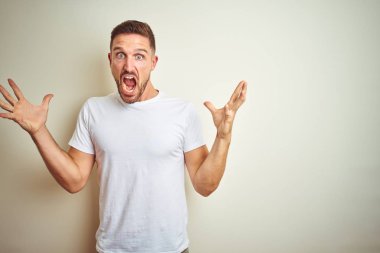 Üzerinde rahat beyaz t-shirt giyen genç yakışıklı adam deli kutluyor arka plan izole ve başarı için kaldırdı kollarını açarak hayrete ve heyecanlı çığlık gözleri açık. Kazanan kavramı