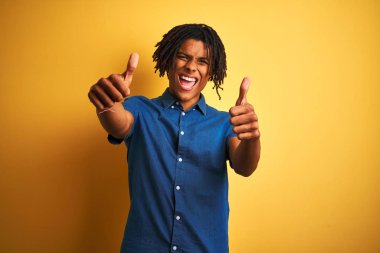 Dreadlocks rahat kot gömlek giyen afro adam izole sarı arka plan üzerinde el ile olumlu jest yapıyor onaylayan ayakta, başparmak kadar gülümseyerek ve başarı için mutlu. Kazanan jest.