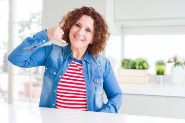 Orta yaşlı, kıvırcık saçlı, kot ceket giyen, gülümseyen, telefonla el ve parmak hareketi yapan, telefonda konuşan bir kadın. İletişim kavramları.