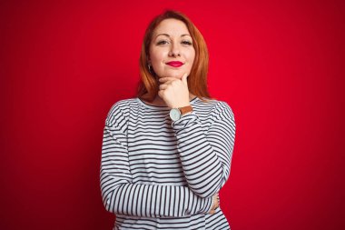 Kızıl saçlı genç bir kadın donanma gömleği giyiyor. Kırmızı izole edilmiş arka plan üzerinde duruyor. Kameraya kendinden emin bir şekilde bakıyor. Olumlu düşünüyorum..