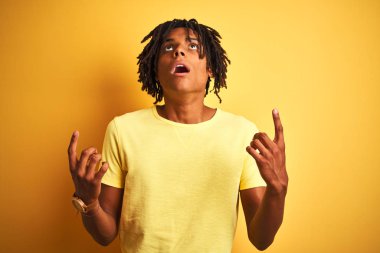 Izole sarı arka plan üzerinde duran rahat t-shirt giyen dreadlocks ile Afro adam şaşırttı ve yukarı bakarak ve parmakları ile işaret ve kolları kaldırdı şaşırttı.