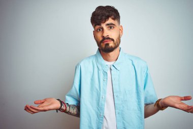 Mavi gömlek giyen dövme giyen genç adam izole beyaz arka plan üzerinde duran habersiz ve kolları ve elleri ile karışık ifade kaldırdı. Şüphe kavramı.