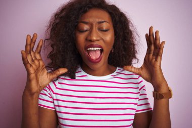 Çizgili tişört giyen genç Afro-Amerikalı kadın izole edilmiş pembe arka planda duruyor ve başarıya deli ve deli gibi seviniyor kollarını kaldırmış ve gözlerini kapamış heyecanla haykırıyor. Kazanan konsept