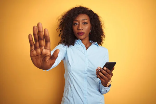 Young african american woman using smartphone standing over isolated yellow background with open hand doing stop sign with serious and confident expression, defense gesture