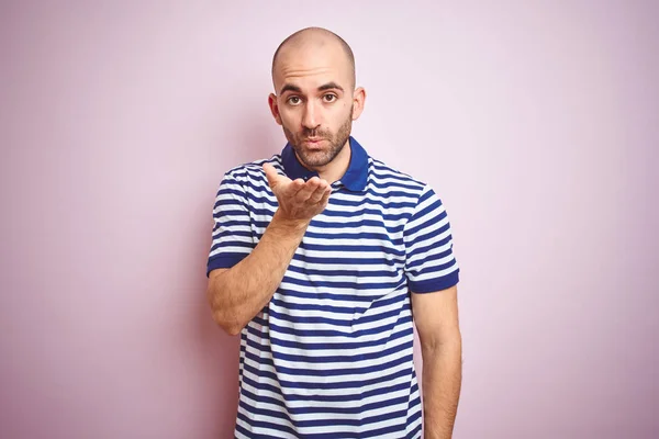 Young bald man with beard wearing casual striped blue t-shirt over pink isolated background looking at the camera blowing a kiss with hand on air being lovely and sexy. Love expression.