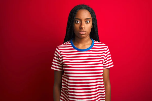 孤立した赤い背景の上に縞模様のTシャツを着た若いアフリカ系アメリカ人女性が顔に真剣な表情でリラックスしています シンプルで自然なカメラを見て — ストック写真