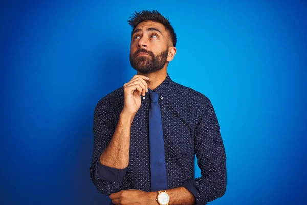 Young indian businessman wearing elegant shirt and tie standing over isolated blue background with hand on chin thinking about question, pensive expression. Smiling with thoughtful face. Doubt concept.
