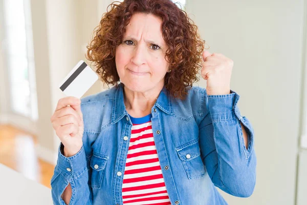 Senior woman holding credit card as payment annoyed and frustrated shouting with anger, crazy and yelling with raised hand, anger concept