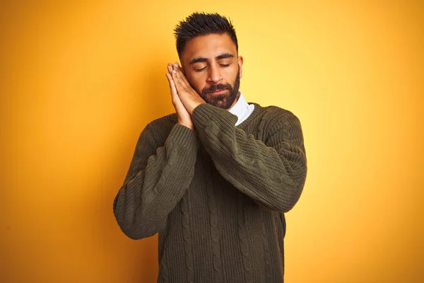 Young indian man wearing green sweater and shirt standing over isolated yellow background sleeping tired dreaming and posing with hands together while smiling with closed eyes.