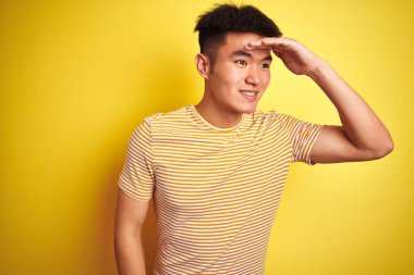Genç asyalı Çinli adam izole sarı arka plan üzerinde duran t-shirt giyen çok mutlu ve başının üzerinde el ile uzak görünümlü gülümsüyor. Arama konsepti.