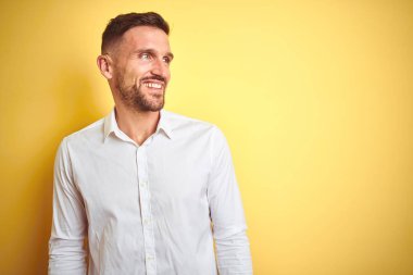 Genç yakışıklı adam sarı izole arka plan üzerinde zarif beyaz gömlek giyen yüzünde gülümseme, doğal ifade ile yan bakıyor. Gülmekten emin.